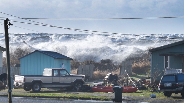 Two homes and two cars appear on a street with huge waves pounding a wall behind them.